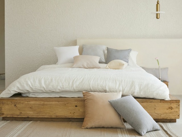 A low wooden bed with white linen and soft neutral cushions