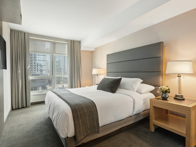 A hotel room with a made king bed, white pillows and a grey bed runner